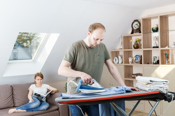 Obraz premium Young woman sitting at the sofa reading the book while her husband is ironing shirt at home.