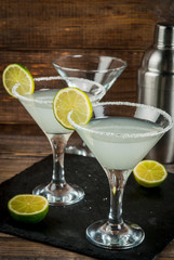 Alcoholic cocktail - gin tonic, martini, margarita. Decorated with slice of lime, with a shaker on a black slate on a wooden table, copy space, vertical