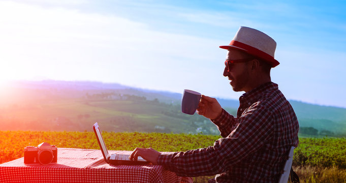 Creative Man Drinking Coffee And Working At Laptop At Sunset On Countryside Background - Silhouette Of Middle Aged Happy Farmer Relaxing With Cup And Pc At Dusk Light  