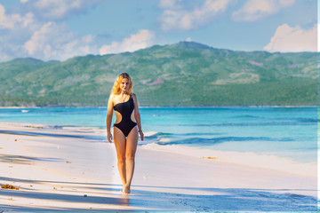 young beautiful girl in black swimwear in full growth walking on the blue beach