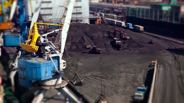 Port Station. Big Pile of Coal on Ground. Excavators Working, Moving Loading. Cars Staying on Rail Road. Cranes on Left. White Building. Wide Shot