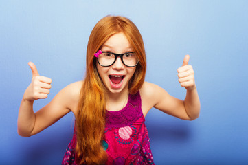 Studio shot of young preteen 9-10 year old redhead girl wearing eyeglasses, standing against blue...