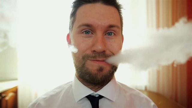 Vape. Young funny white businessman with beard and mustache admits puffs of steam from the electronic cigarette in the kitchen. 
