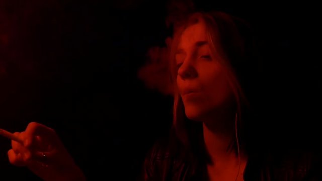 Pretty nice girl and smokes a cigarette in club exhailing smoke. Red light glow and smoke breathing. Sad girl waiting in the bar. Smoking area. 50 fps.