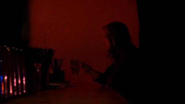 Pretty nice girl in restaurant drinks from a whiskey glass. Bar stand with drinks, menu, sign, glass. Red light and glow. 50 fps