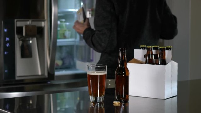 Man in modern kitchen with beer in glass on countertop reaches into the refrigerator to get a snack of cheese
