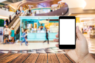 Mock up image of hand holding black mobile phone with blank white screen, Empty wood table top on blurred images shopping mall for background, perspective wood can be used for display products.