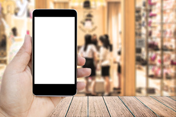 Mock up image of hand holding black mobile phone with blank white screen, Empty wood table top on blurred images shopping mall for background, perspective wood can be used for display products.