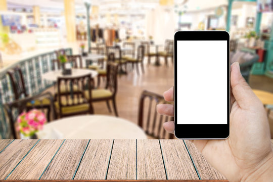 Mock up image of hand holding black mobile phone with blank white screen, Empty wood table top on blurred images restaurant for background, perspective wood can be used for display products.