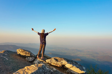 Young traveler is standing at the mountain view point, rises her hand up under the beautiful clear sky.  