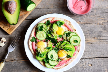 Tortilla piled high with fried quail eggs, avocado, cucumbers, sesame and parsley and lathered with beetroot hummus. Half avocado, beetroot hummus in bowl, fork on vintage wooden background. Top view