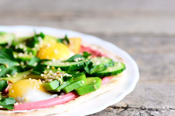 Healthy tortilla on a plate and on a wooden table. Simple tortilla with fried quail eggs, avocado, cucumbers, sesame seeds, parsley and beetroot hummus. Delicious rustic breakfast idea. Closeup