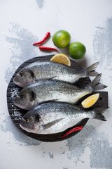 Wooden chopping board with three raw fresh sea breams and seasonings, above view on a white concrete background