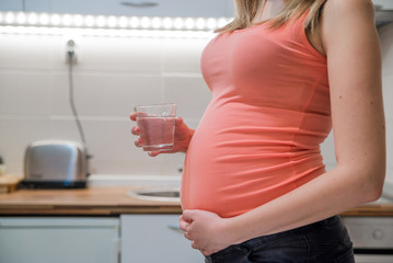 Pregnant woman drinking glass of water at home in the kitchen. Happy pregnant woman holding a glass of water while standing in the kitchen