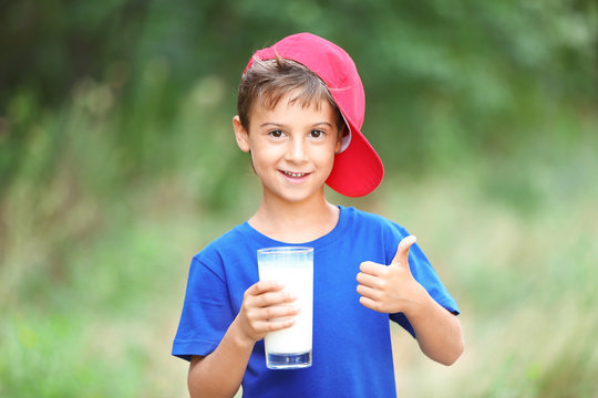 Cheerful kid in blue shirt and red hat holding glass of milk on blurred background