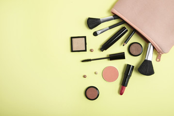 Make up products spilling out of a pastel pink cosmetics bag on to a pale yellow background, with...