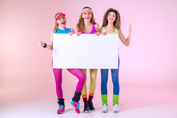 Fototapeta premium cheerful sporty women holding blank banner on pink