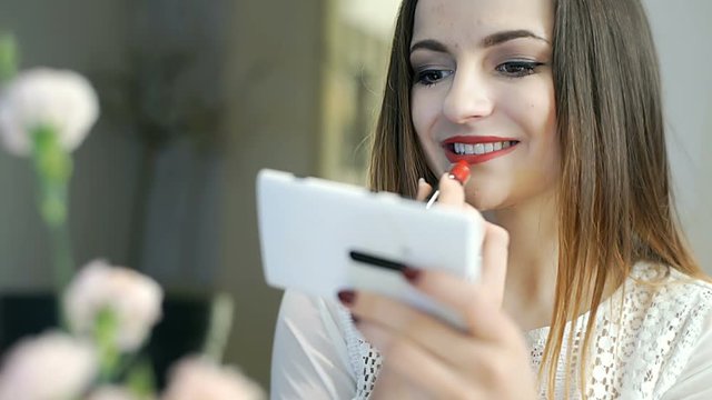 Romantic girl using smartphone as a mirror while painting lips with red lipstick
