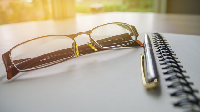 Stock picture of a silver pen and open spiral notebook lying on a dark brown wooden desk with eyeglasses in the background.