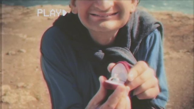 Fake VHS tape: an ugly man kneels in front of her girlfriend at a romantic place and shows an engagement ring. Point-of-view of the woman. Funny scene.

