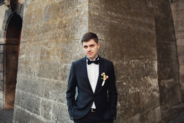 Fototapeta na wymiar Handsome brunette groom in tuxedo with white boutonniere stands on the corner of stone tower