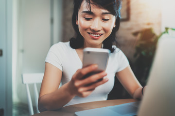 Young smiling Asian woman spending rest time at home and using smartphone for texting with friends.Selective focus. Blurred background, flares effect.