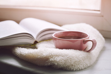 Cozy home still life: cup of hot coffee and opened book with warm plaid on windowsill against snow. Free copy space