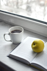 Cozy home still life: cup of hot coffee and opened book with green apple on windowsill. Free copy space