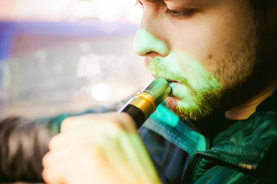 young vaper man with beard vaping mechanical mod. Guy smokes an electronic cigarette by blowing a smoke vapor. Bringing rda mouthpiece to the lips on colorful background