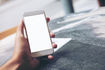 Mockup image of hand holding white mobile phone with blank white screen on marble table in cafe