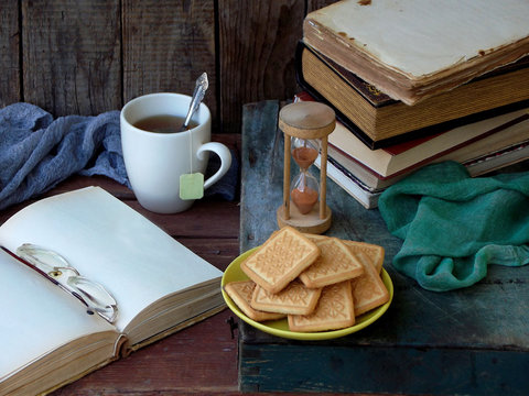 The composition of a stack of old books, open book, tea cups, glasses and plates of sugar cookies on a wooden background. Vintage.