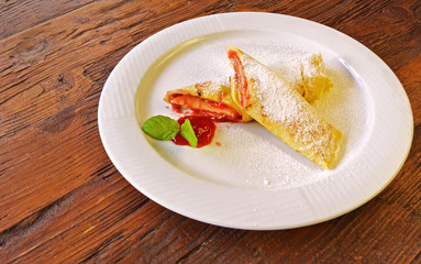 Rolled pancakes with powdered sugar and strawberry jam. French crepes on white plate on wooden background