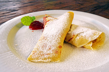 Rolled pancakes with powdered sugar and strawberry jam. French crepes on white plate on wooden background