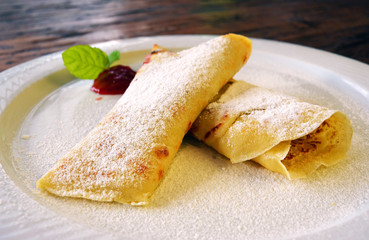 Rolled pancakes with powdered sugar and strawberry jam. French crepes on white plate on wooden background