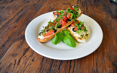 Preparing delicious Italian tomato bruschetta with chopped vegetables, olives, cheese and oil on grilled or toasted crusty baguette sprinkled with seasoning and spices on a plate on wooden background