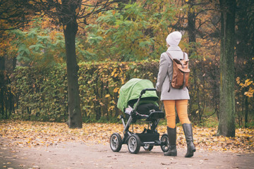 Young mother pushing stroller in autumn park. Young woman strolling with baby in green buggy outdoors