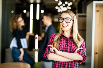 Portrait of a positive looking young woman standing with her arms crossed with coworkers talking in the background.