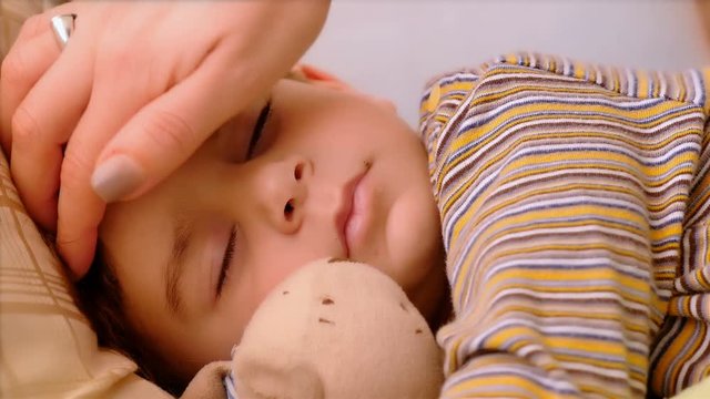Portrait of 3 years old boy sleeping with teddy bear. His mother hand caressing him.