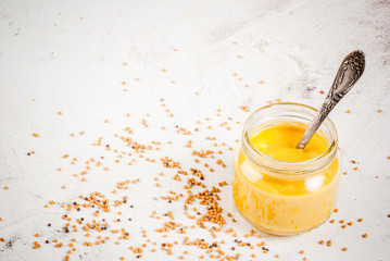 Fresh yellow American mustard in a small jar with a spoon. With mustard grains, on a white stone background, copy space