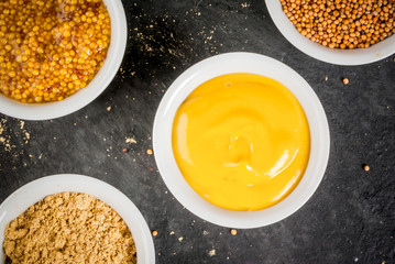 Selection of 4 kinds of mustard: American yellow, French with whole grains, dry grain and ground mustard. In white saucers, on a white stone table, close top view, copy space