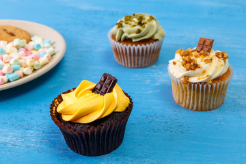 Cupcakes with buttercream. Sweet cake with pistachio nuts cream and small chocolate bar. Colored marshmallows. Dessert with topping. On blue background.
