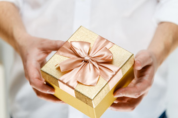 Male hands holding a gift box. Present wrapped with ribbon and bow. Christmas or birthday golden paper package. Man in white shirt.