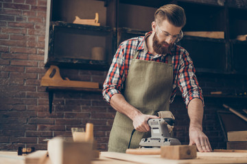 Photo of young handsome serious handicraftsman with safety glasses using grinder and sandpaper in...