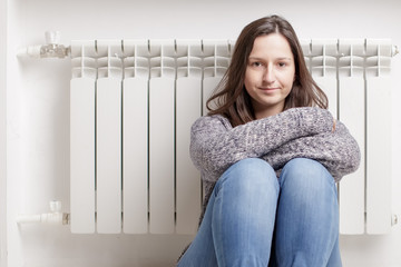 Young beautiful girl froze and warms hands near a radiator, dressed in a sweater 
