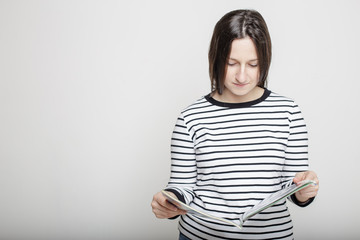 Fototapeta premium Young attractive woman carefully reading a magazine in their hands on a blank gray wall background in a striped sweater