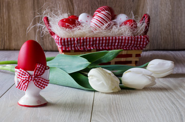 Red Easter egg in egg cup with bow on wooden table top. Easter eggs in basket, white tulips.