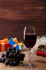 Red wine bottle. Wineglass with dark grapes branch. Gift box with bow on rustic wooden background.