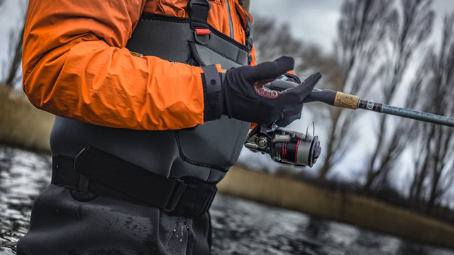 Angler h&auml;lt Spinnrute im Wasser stehend nahe Ufer mit B&auml;umen in Wathose und Handschuhen und kurbelt Rolle