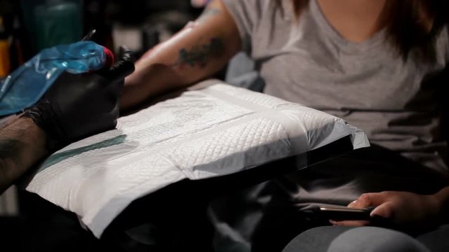 Professional tattoo artist makes a tattoo on a young girl's hand. Girl is watching something in the phone