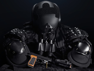 Black military tactical gun, helmet, gloves, cartridge belt, bandolier, gun shell, knife, binocular and knee protection laying on a black table.